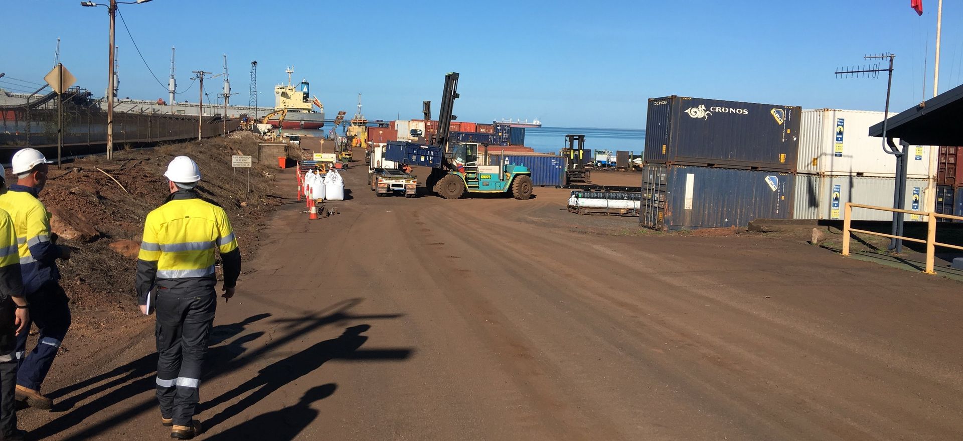 Workers wearing safety gear walk along a port with shipping containers and cranes.