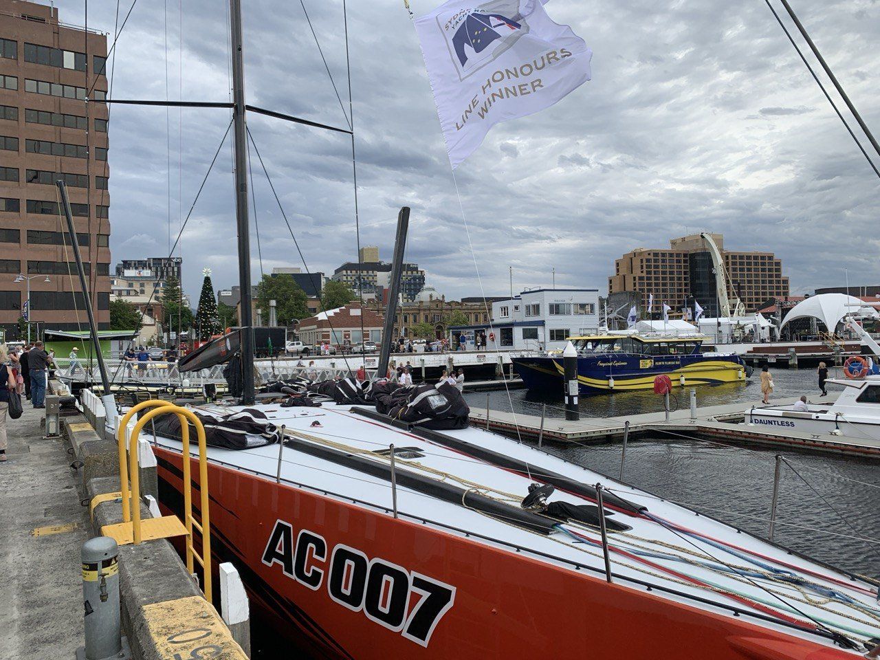 A marina scene with boats docked, including a large racing yacht with "AC007" on the side.