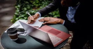 Person writing notes beside laptop outdoors.